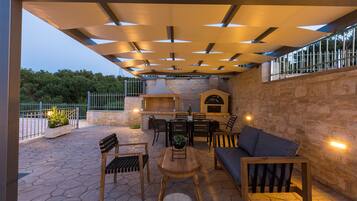 Un patio al aire libre con suelo de piedra, mobiliario de madera, una parrilla y un muro de piedra.