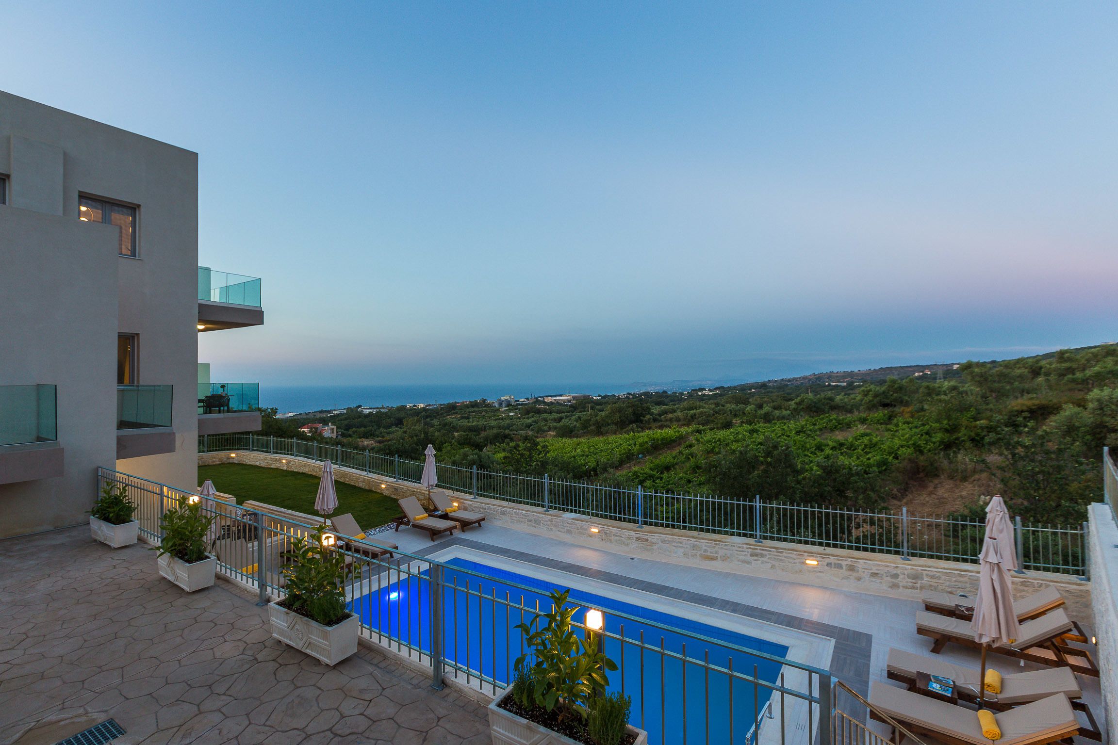 A rooftop pool with lounge chairs and umbrellas, overlooking a scenic view of the sea and hills.