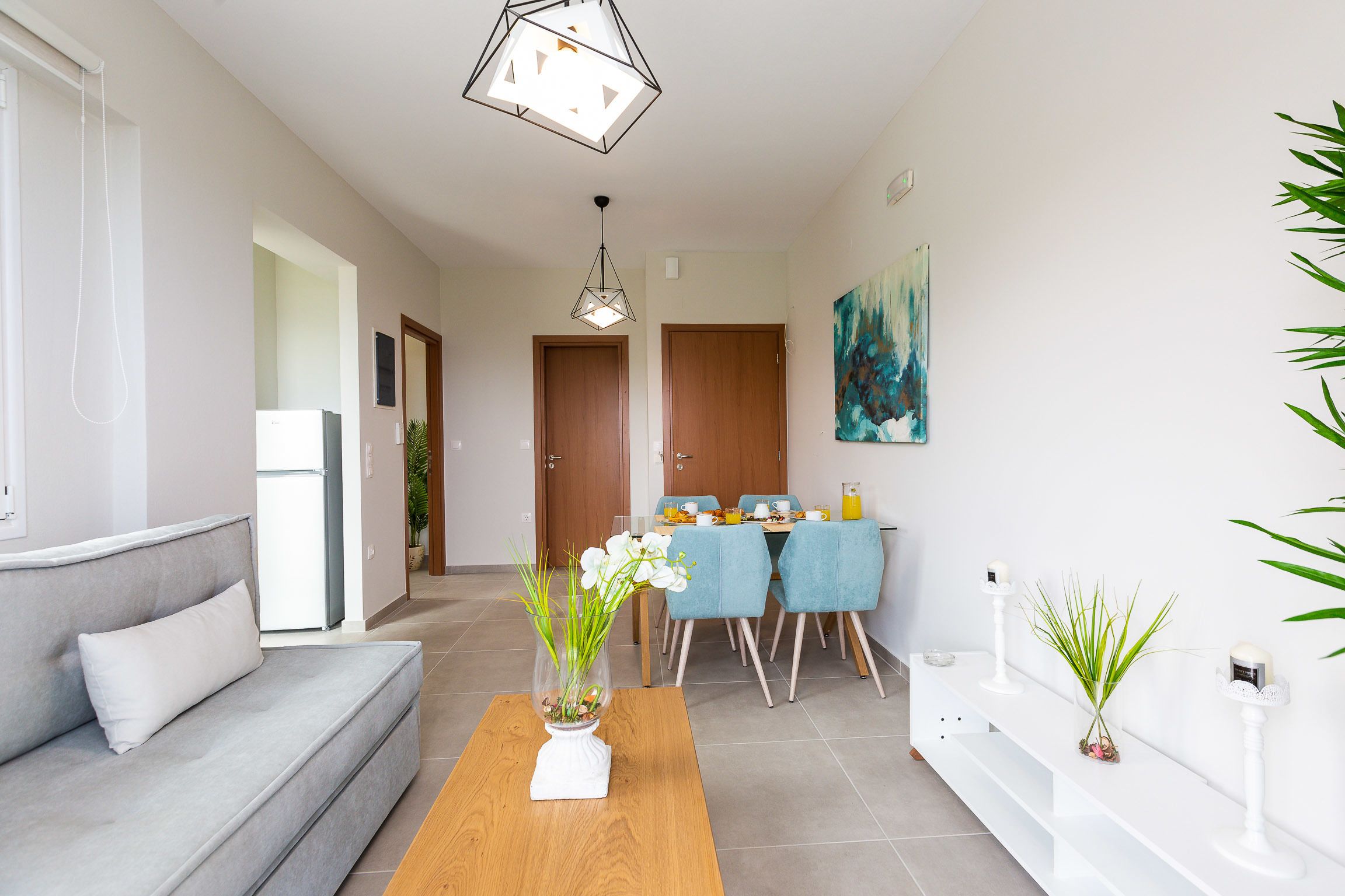 A modern living room with a grey sofa, a wooden coffee table, and a dining area with blue chairs and a wooden table.