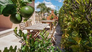 Een terras buiten met houten tafels en stoelen, omringd door weelderig groen en een heldere blauwe lucht.