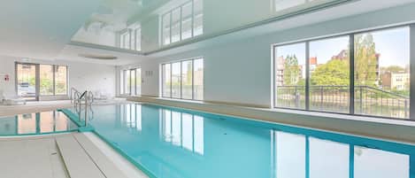 Una piscina interior con paredes de vidrio transparente, suelo de baldosas y vistas a edificios exteriores.