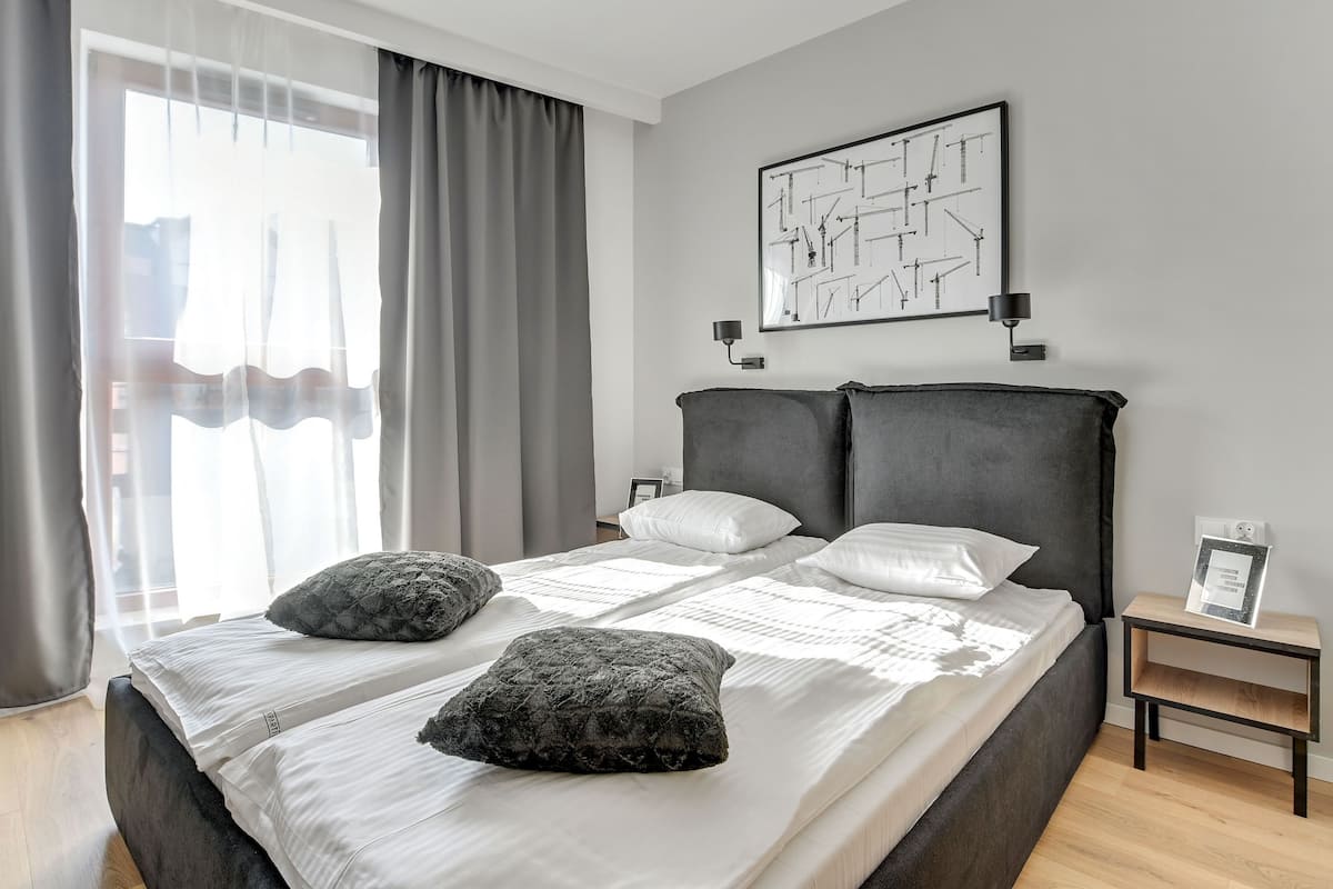 Ein modernes Schlafzimmer mit einem großen Bett, grauen Vorhängen, einem an der Wand hängenden abstrakten Kunstwerk und einem Nachttisch mit einer Lampe.