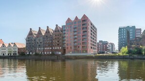 A riverside view of a city with historic and modern buildings, including a prominent red building with a unique architectural design. - Apartinfo Apartments Waterlane Island (Gdansk)