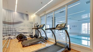 A gym with treadmills, a rowing machine, and a large window overlooking a pool. - Apartinfo Apartments Waterlane Island (Gdansk)