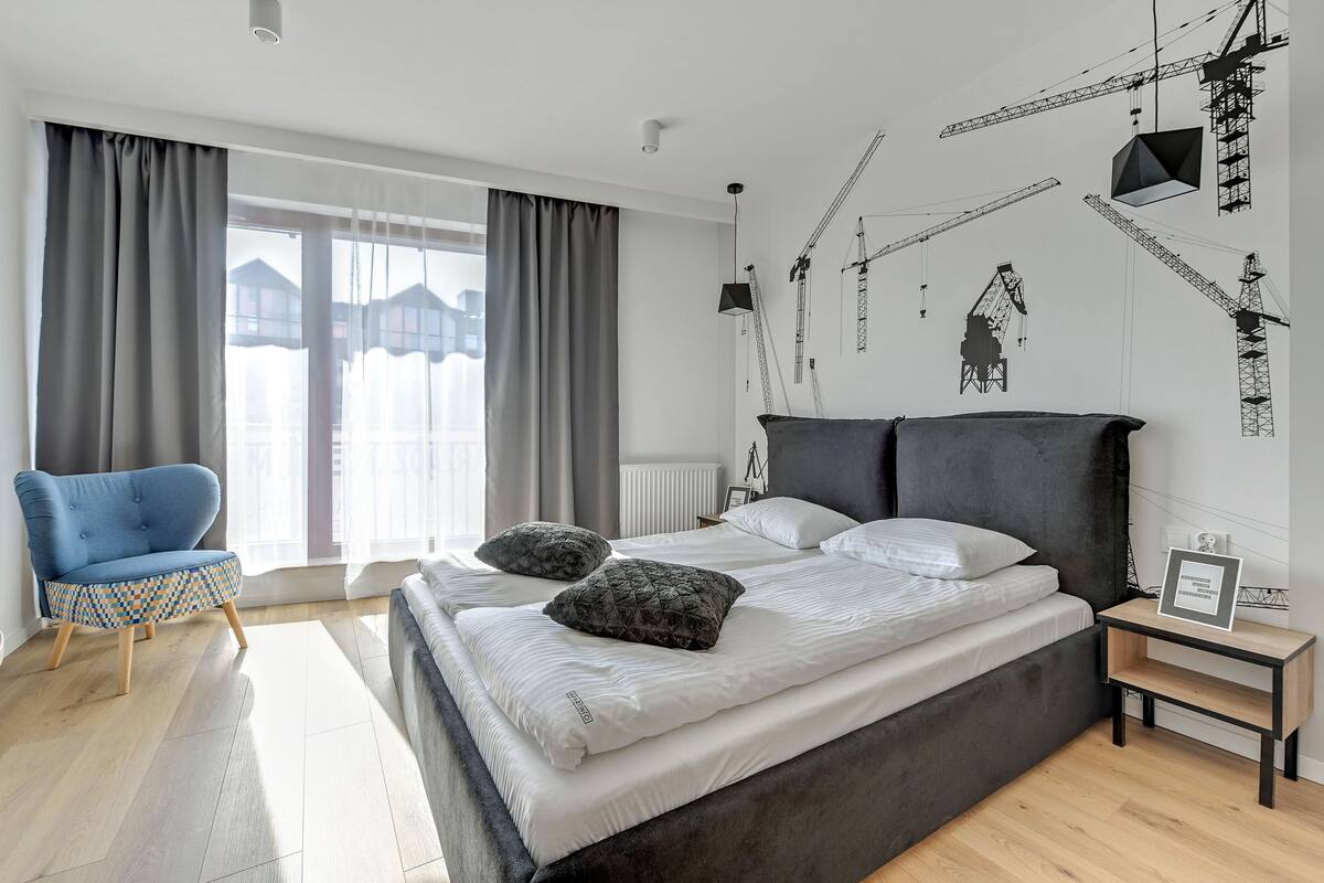 Ein modernes Schlafzimmer mit einem großen Bett, einem blauen Sessel und einer Wand mit Illustrationen von Baukränen.