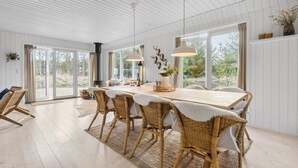 Un comedor amplio con una larga mesa de madera, sillas de mimbre y suaves mantas de piel blanca, y ventanas grandes que ofrecen vistas al exterior.