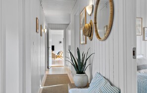 A hallway with white paneled walls, a potted plant, a striped pillow, and a woven basket on a woven stool.