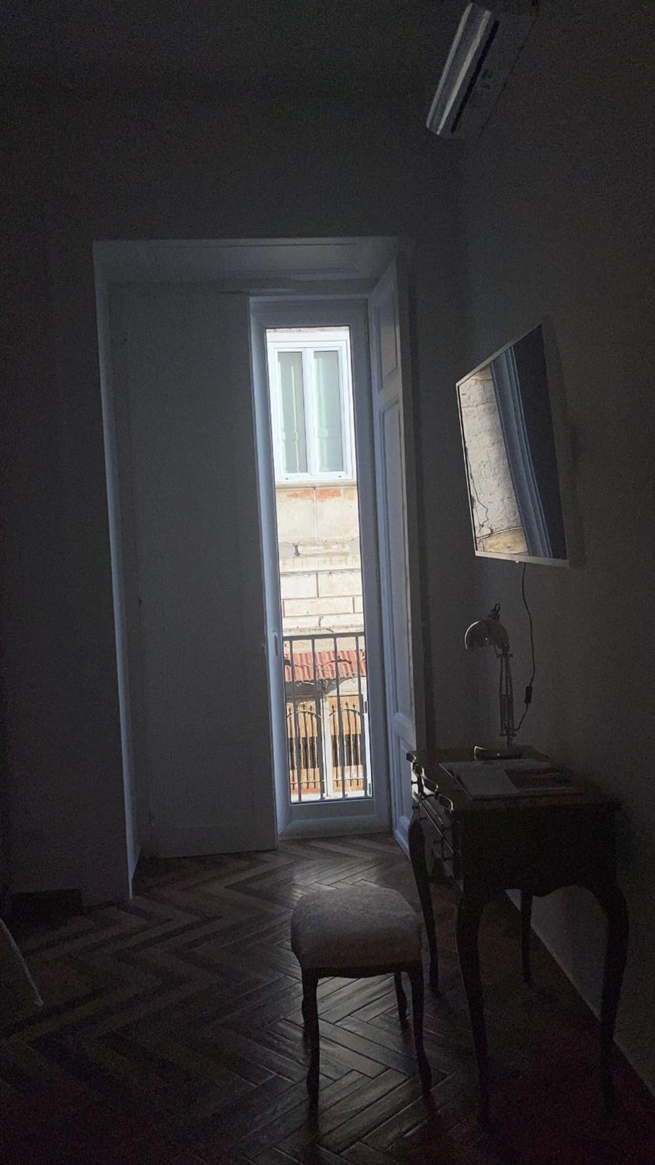 Ein schwach beleuchteter Raum mit Holzboden, einem kleinen Tisch, einem Stuhl und einem Spiegel an der Wand. Eine Tür führt zu einem Balkon.