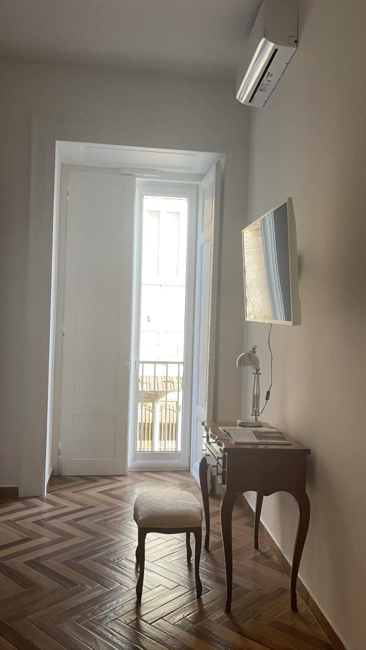 A hallway with a wooden herringbone floor, a white door, a small table with a lamp, and a wall-mounted mirror.