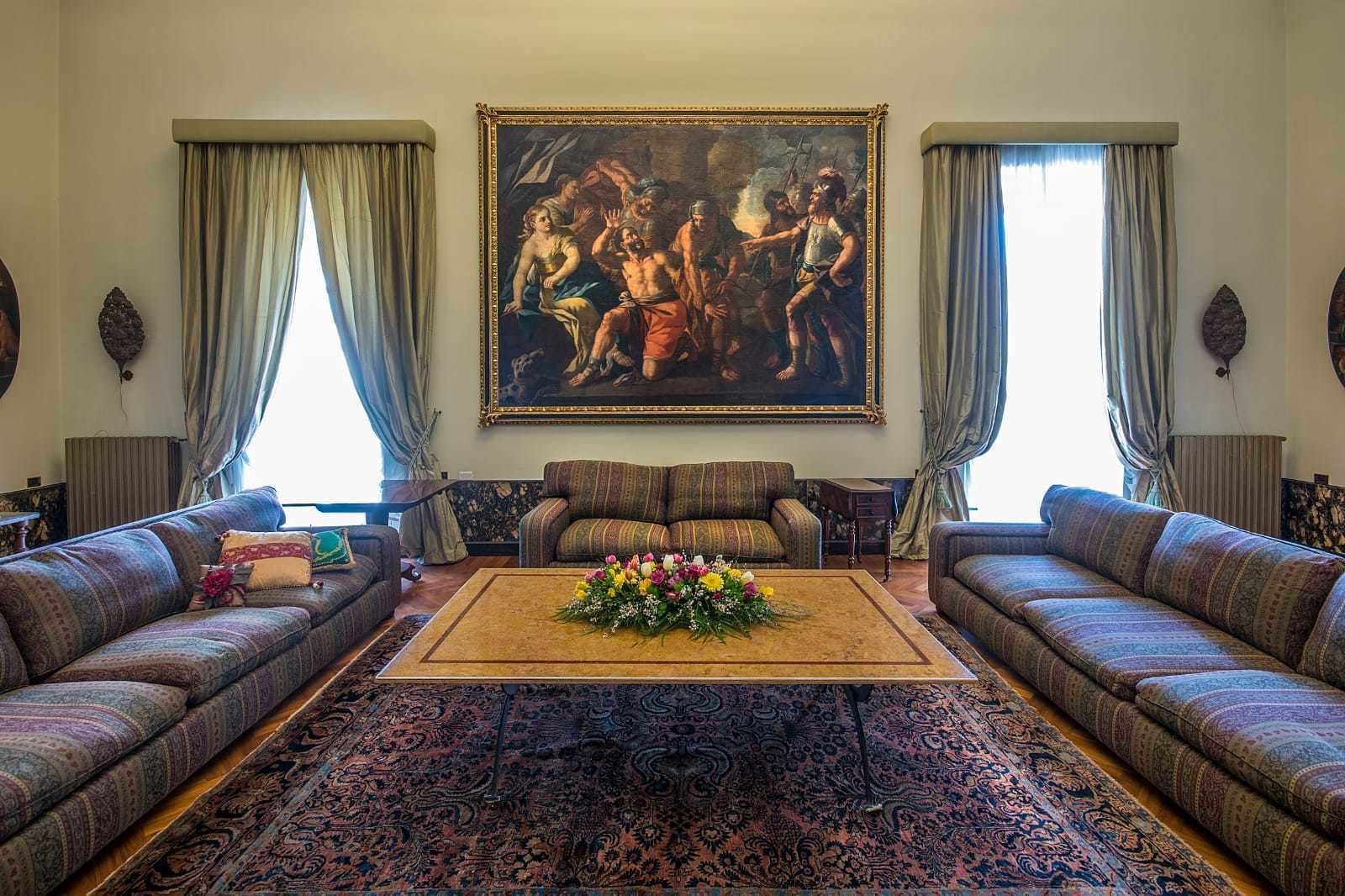 Una sala de estar con un cuadro clásico grande, dos sofás, una mesa de centro con flores y alfombras estampadas.