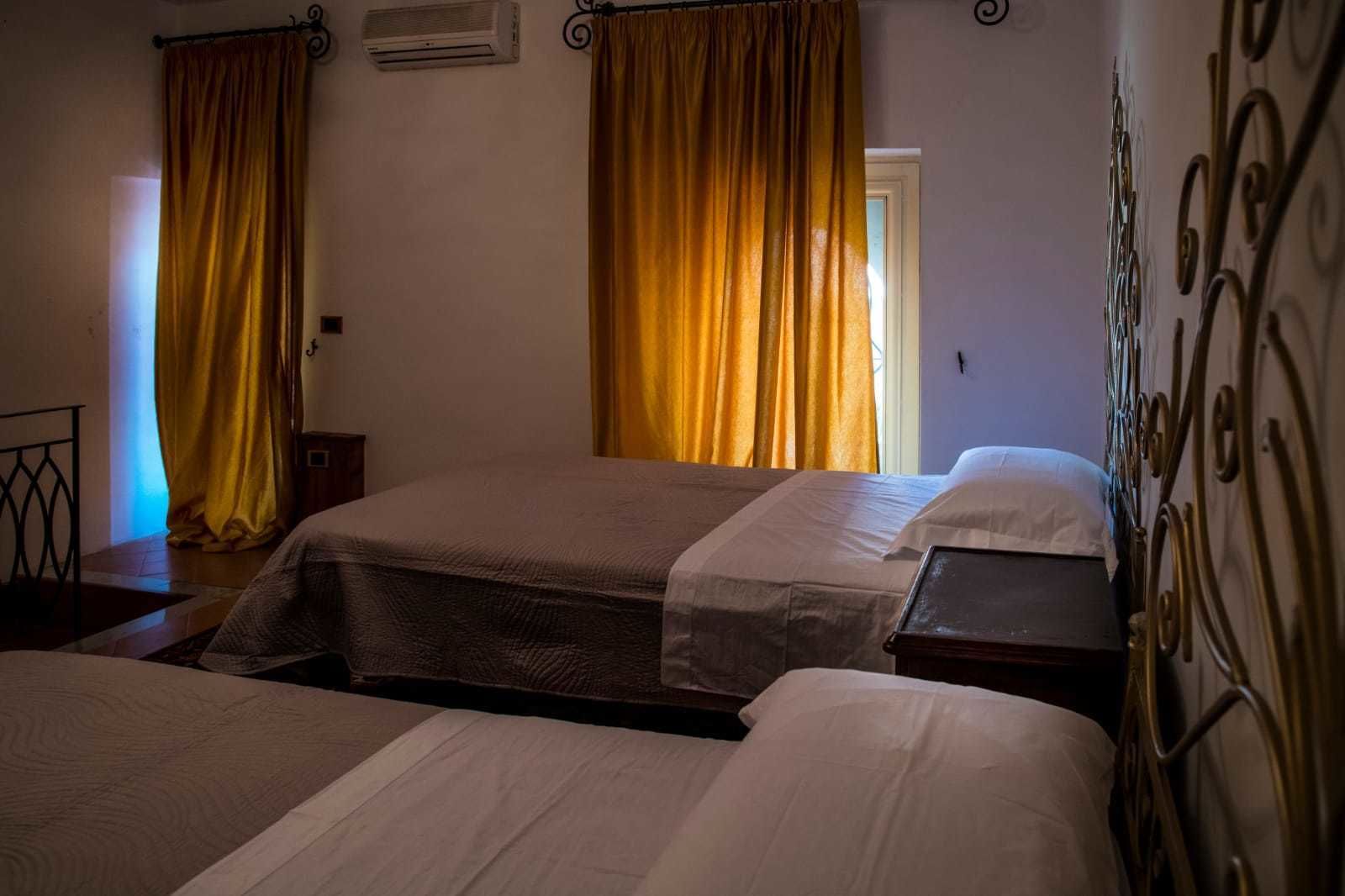 Een hotelkamer met twee bedden, een raam met gele gordijnen en een metalen bedframe.