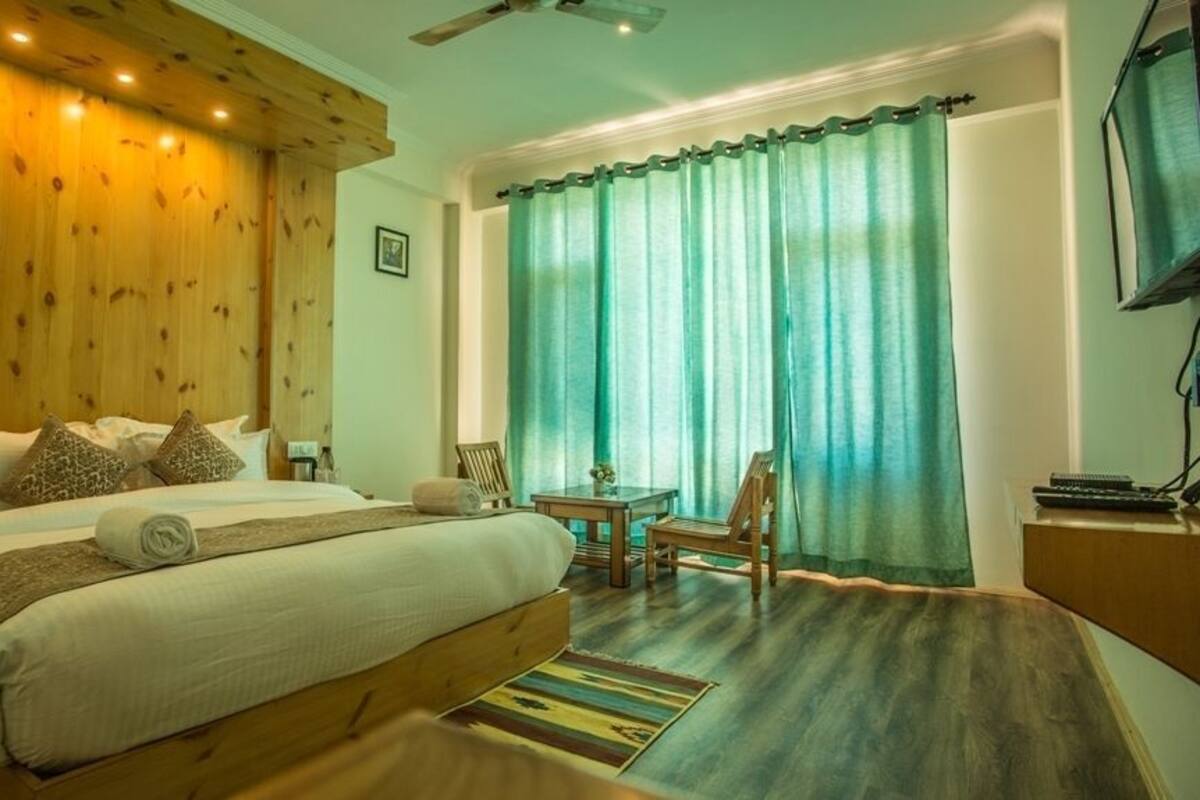 Un dormitorio con cabecera de madera, cama con almohadas, una mesita, una silla y un televisor sobre un mueble de madera.