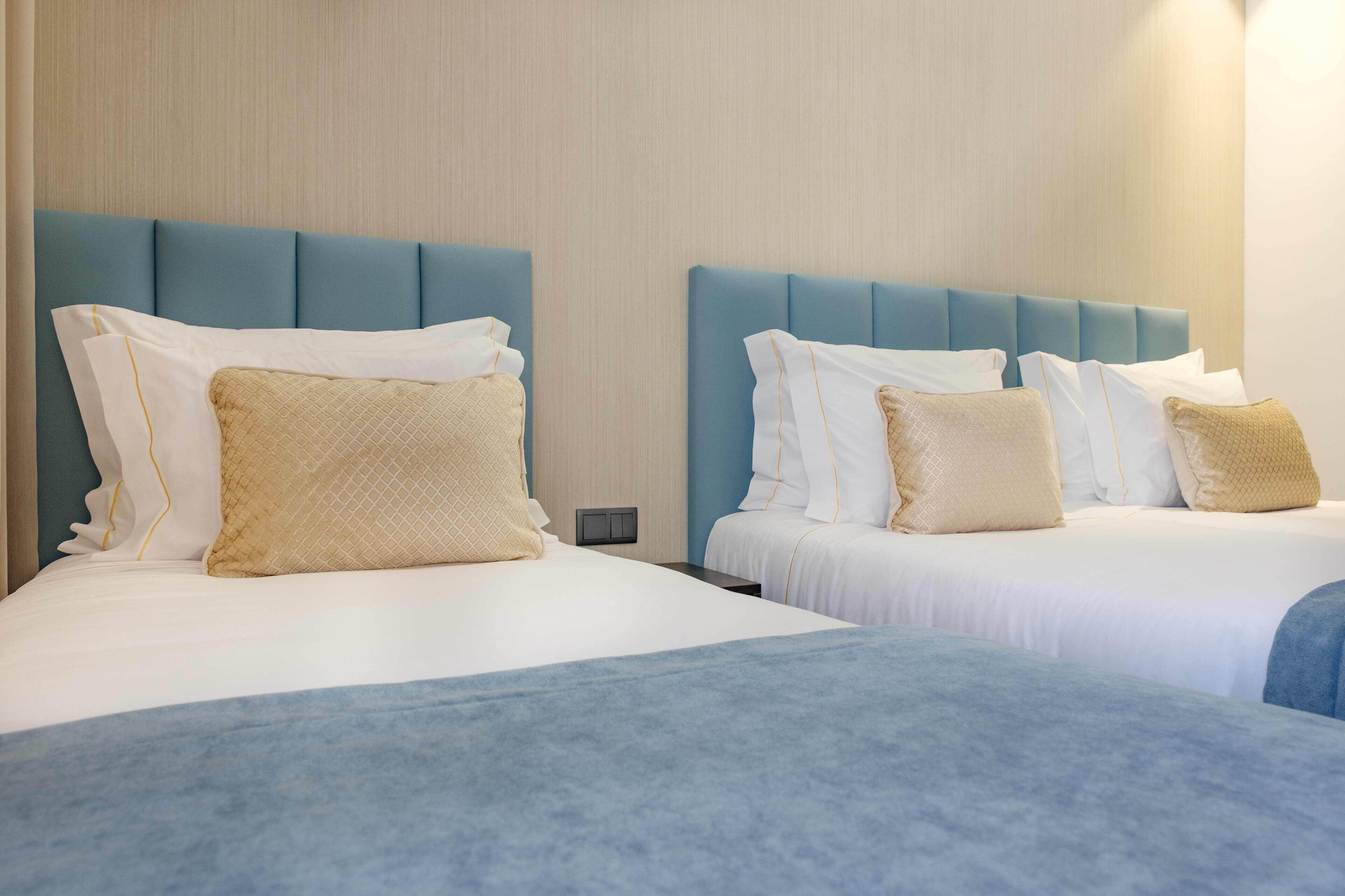 Dos camas con cabeceros azules, ropa de cama blanca y almohadas color beige en una habitación de hotel.