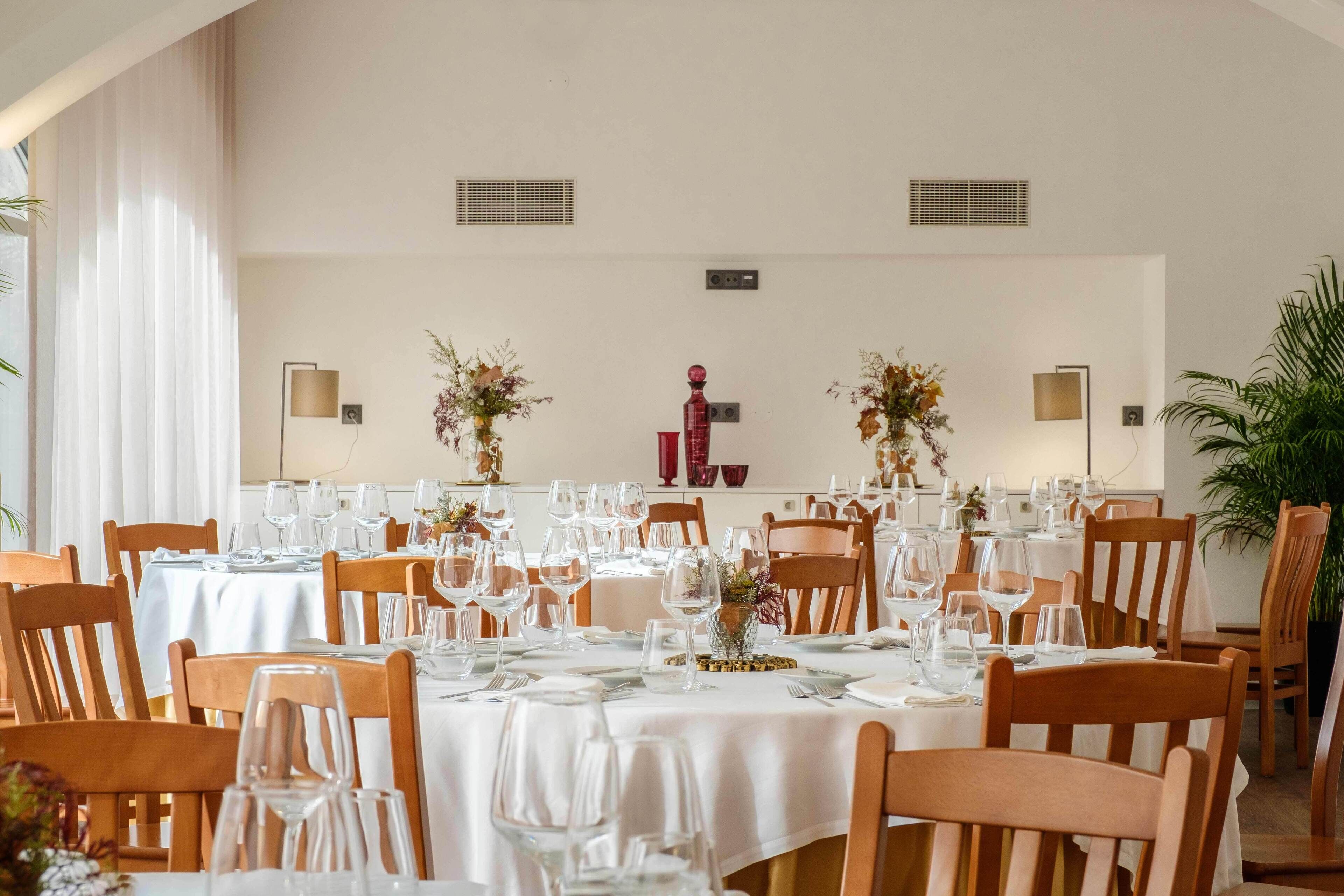 Un comedor con mesas dispuestas para una comida, con manteles blancos, sillas de madera y centros de mesa decorativos.