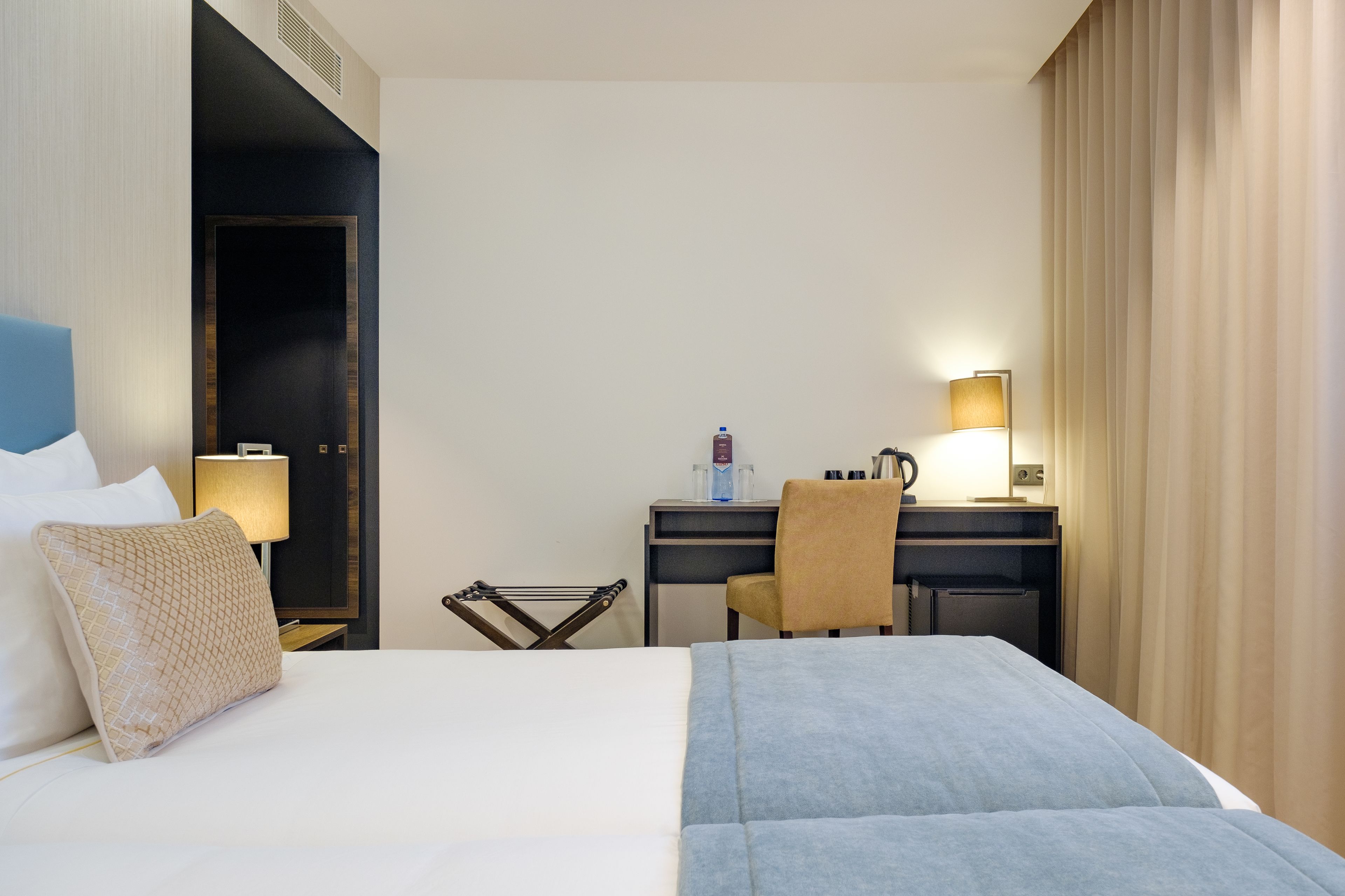 A hotel room with a bed, a desk, a chair, a lamp, and a window with curtains.
