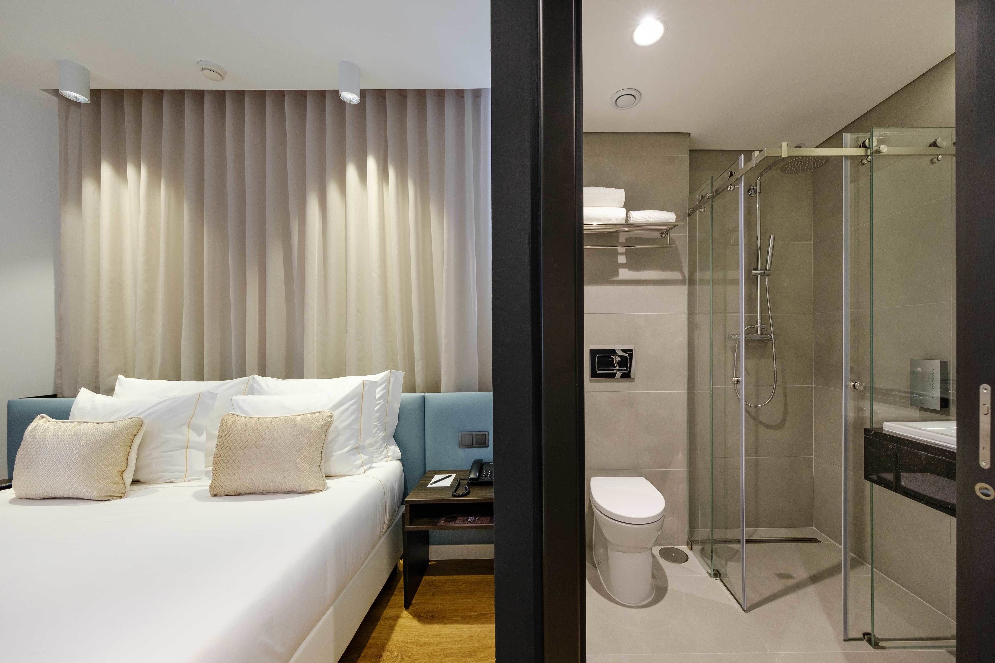 Quarto de hotel moderno com uma cama grande, mesa de cabeceira, escrivaninha, chuveiro com portas de vidro e vaso sanitário.