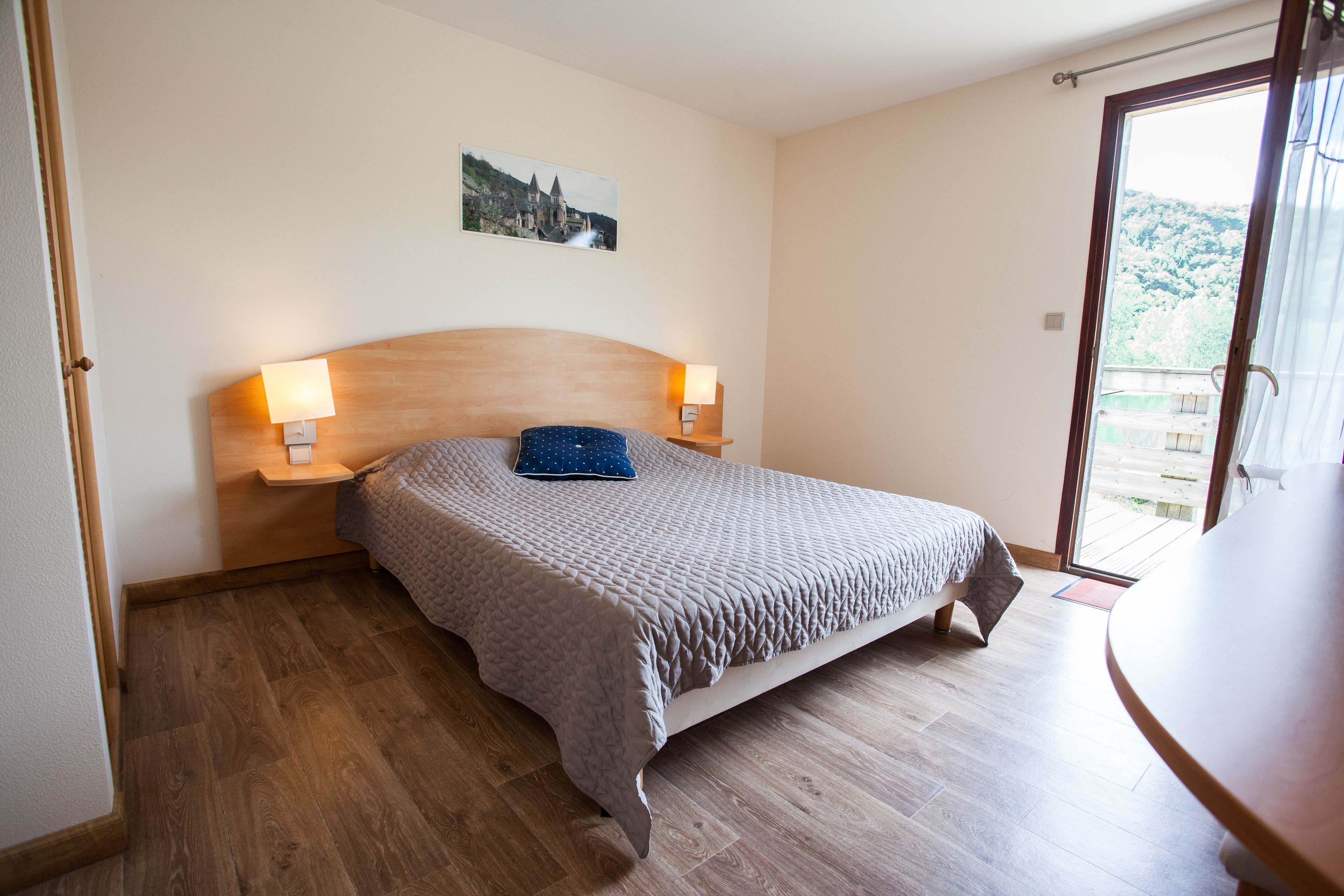 Ein Schlafzimmer mit einem hölzernen Kopfteil, einem Bett mit grauer Bettdecke, zwei Nachttischlampen und einem Bild an der Wand.