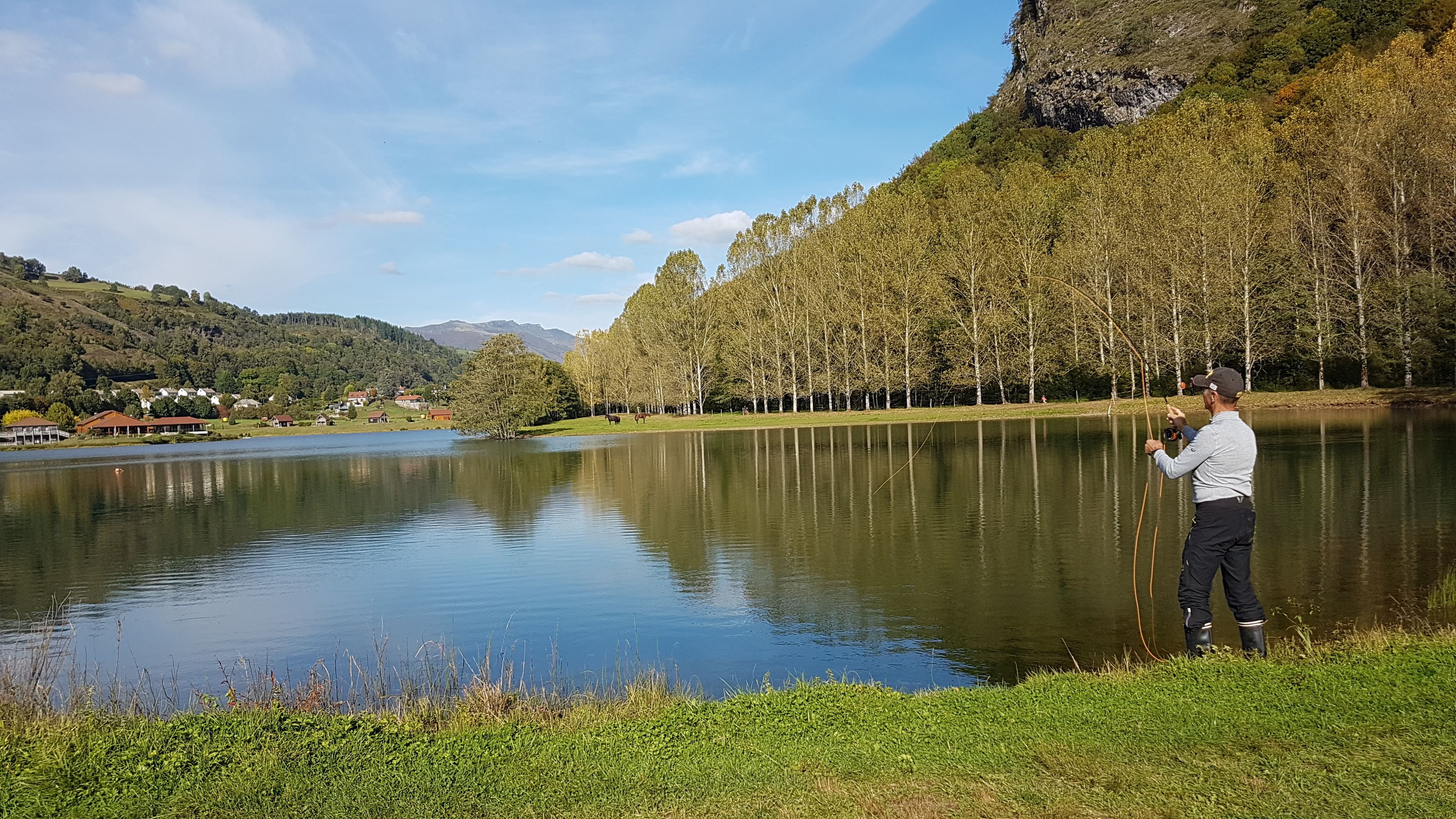 Un lago tranquilo con una persona tomando una fotografía, rodeado de árboles y un pueblo distante.