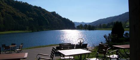 Un patio junto al lago con mesas y sillas, con vista a un lago tranquilo y colinas boscosas.