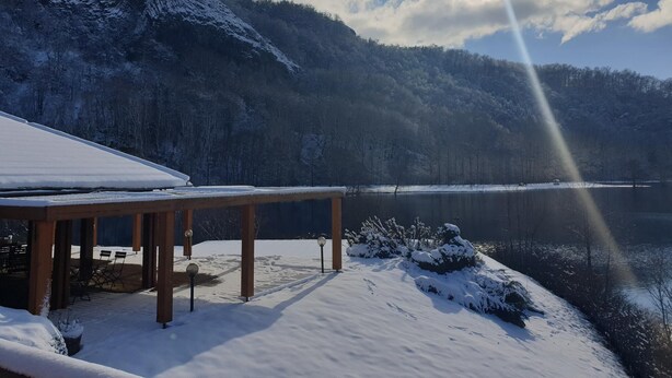 Een houten terras met een sneeuwbedekte dakrand met uitzicht op een meer, omgeven door beboste bergen.