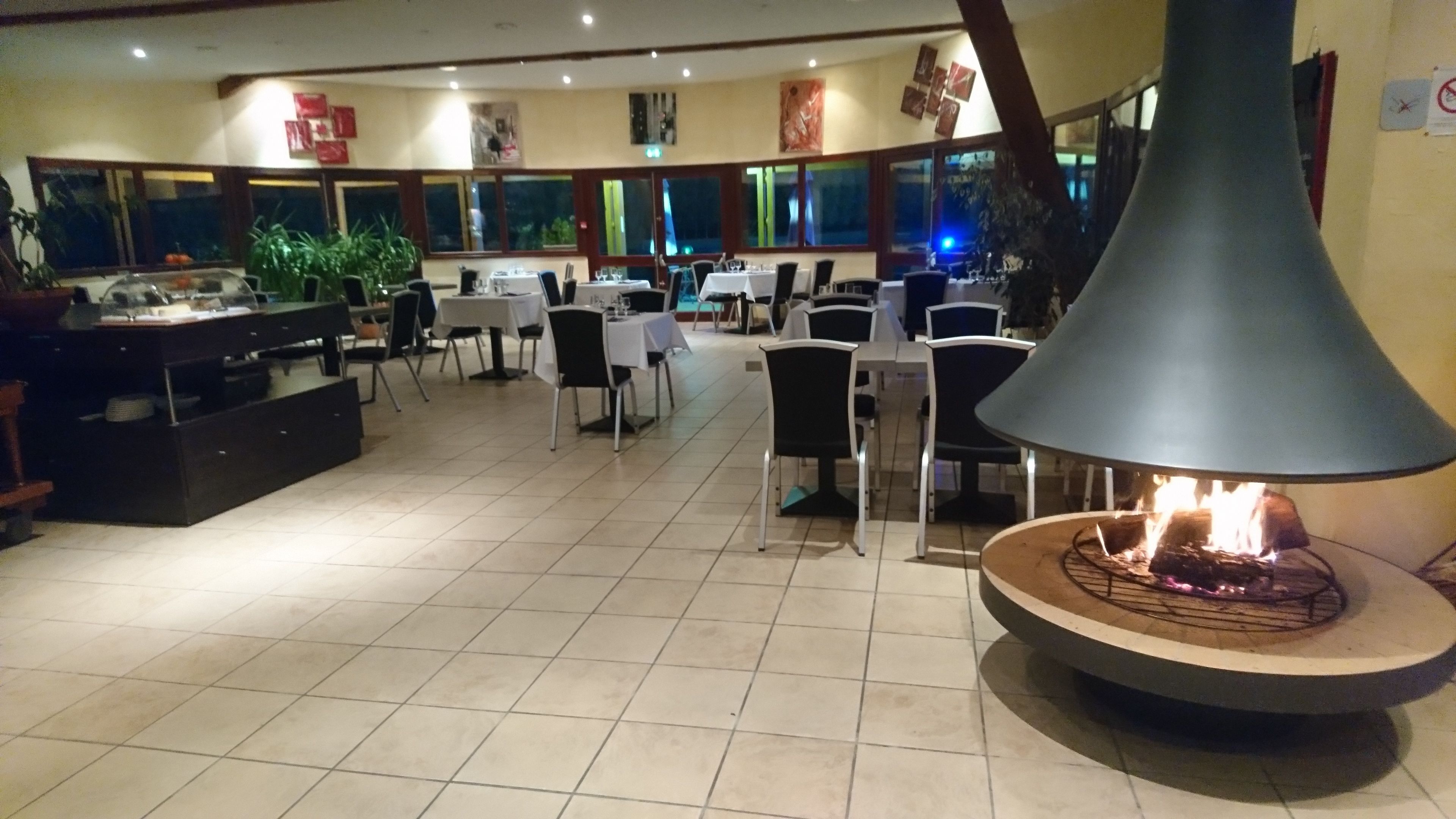 Un restaurante con una chimenea central, varias mesas, sillas y una zona de cocina visible al fondo.