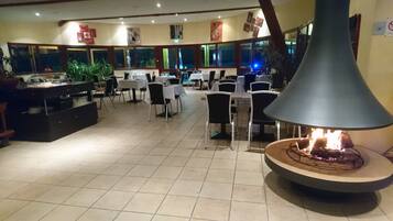 A restaurant with a central fire feature, multiple tables, chairs, and a kitchen area visible in the background.
