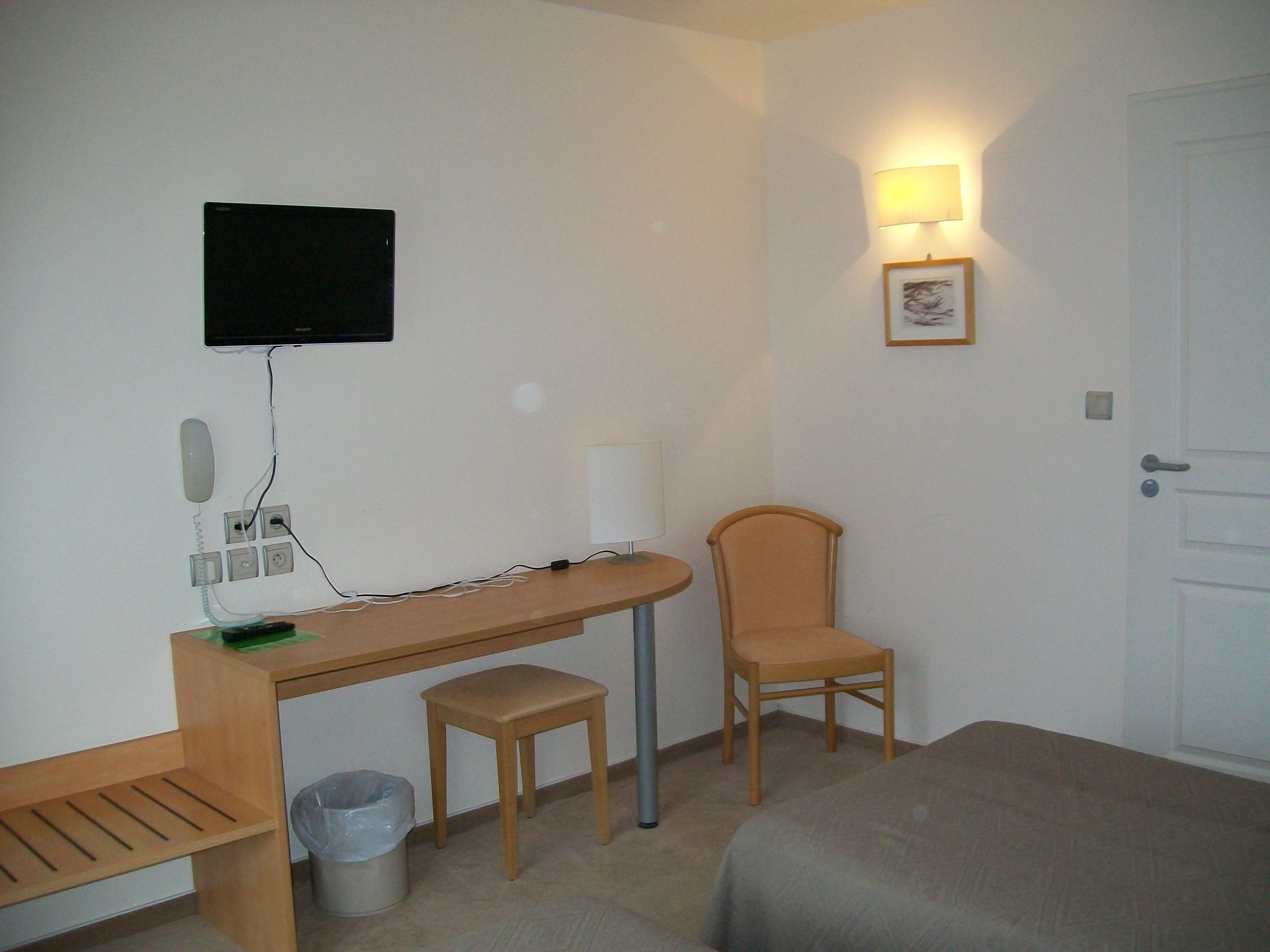 Une chambre d’hôtel avec un bureau en bois, une chaise, un petit tabouret, une poubelle, un lit, un téléviseur fixé au mur et une lampe fixée au mur.