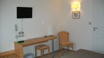 Une chambre d’hôtel avec un bureau en bois, une chaise, un petit tabouret, une poubelle, un lit, un téléviseur fixé au mur et une lampe fixée au mur.