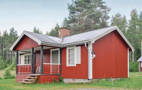 Una cabaña de madera roja con un porche, una chimenea y un pequeño jardín delantero.
