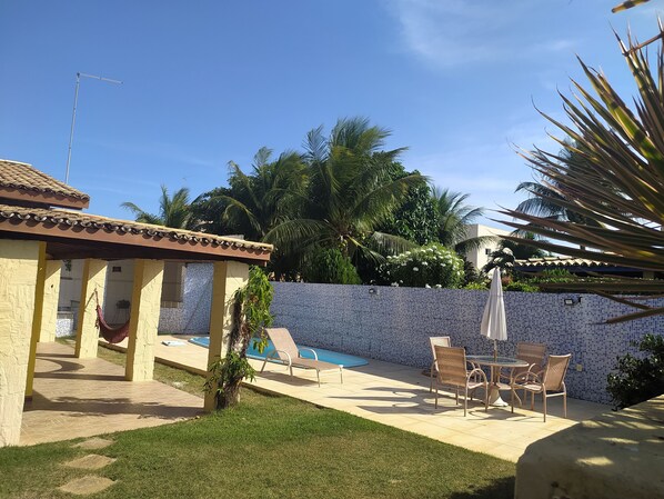 A pool area with a tiled wall, lounge chairs, and a small palm tree. - House in a gated community with a pool and air conditioning in the bedrooms, ideal for families. (Camaçari)