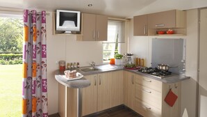 A modern kitchen with wooden cabinets, a gas stove, and a window with blinds. - Mobilhome Camping Arquebuse (Athée)