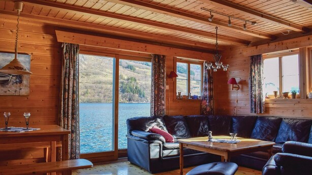 A cozy cabin interior with a wooden ceiling, large windows offering a view of a lake, a leather sofa, a wooden table, and a bench. - 2 bedroom gorgeous home in ølensvåg (Ølensvåg)