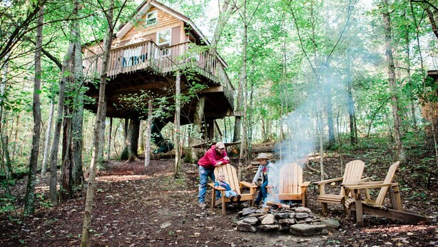 Una cabaña de madera con terraza, rodeada de árboles, y un hoyo para fuego con sillas cerca.