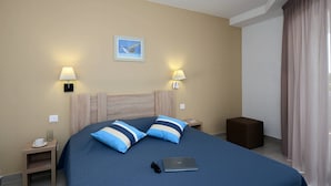 A hotel room with a bed, two bedside lamps, a laptop, a small table with a cup and glasses, and a picture on the wall.