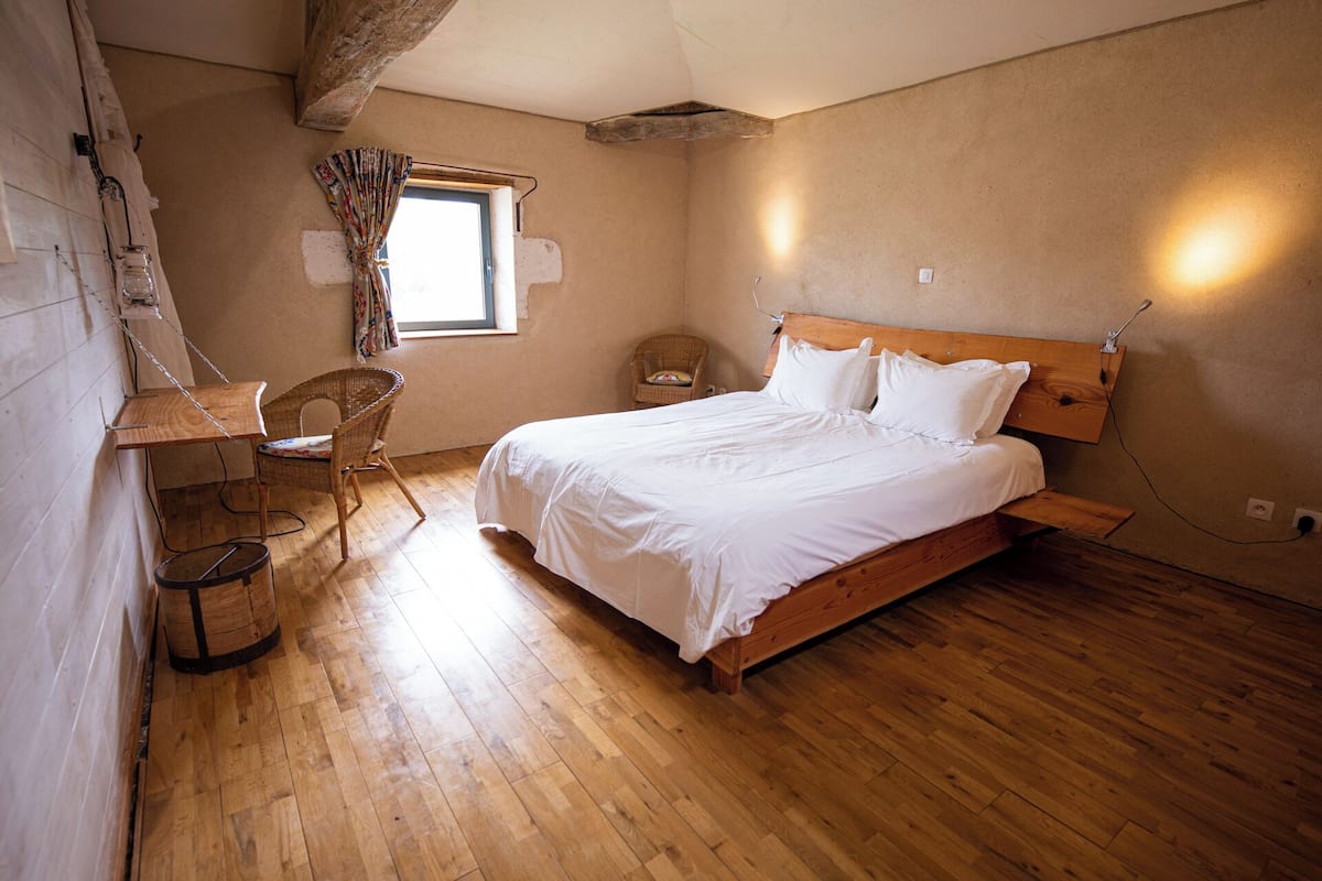 Ein Schlafzimmer mit einem hölzernen Bett, einem Sessel, einem kleinen Tisch und einem Fenster mit Vorhängen.