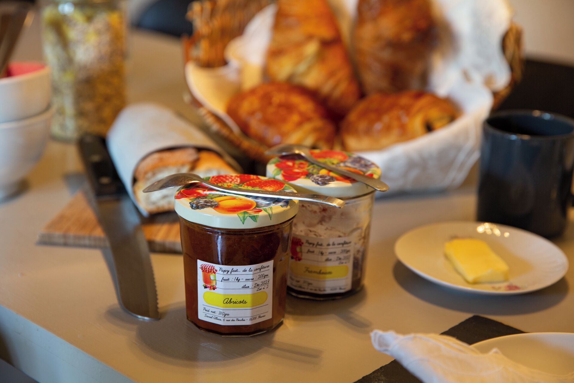 Taula amb pots de conserves, cistella de croissants, plat amb mantega i un ganivet.