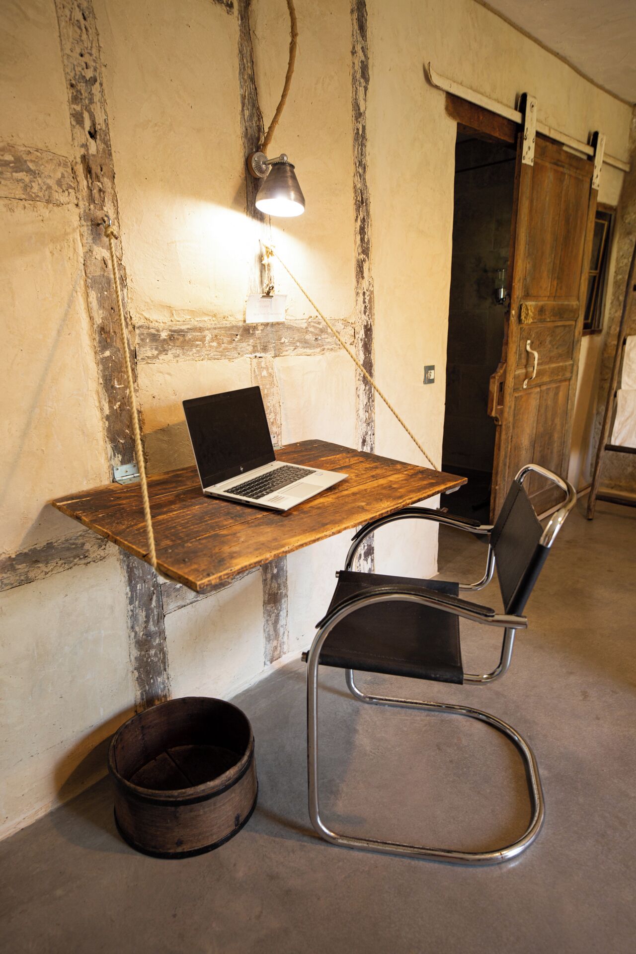 Un espace de travail rustique avec un bureau en bois, un ordinateur portable, une chaise en métal et une lampe suspendue.
