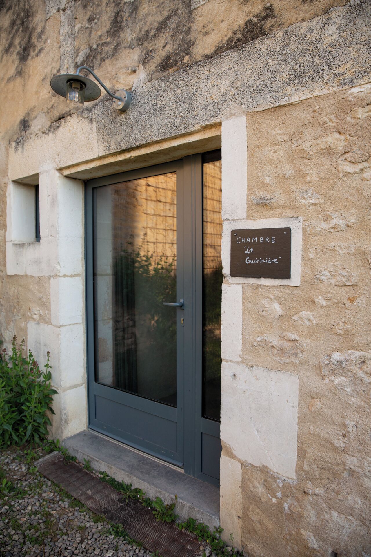 A building entrance with a door, a sign reading "CHAMBRE la GAVERNIERIE," and a small outdoor area with plants.