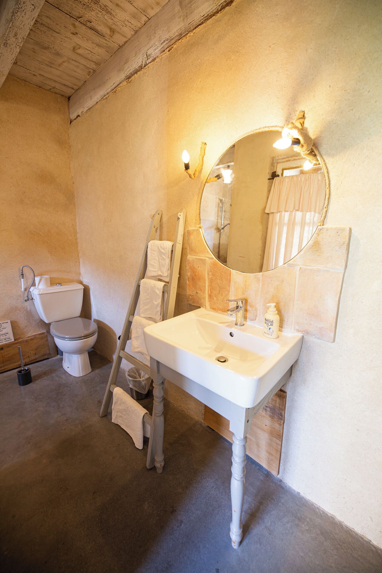 Ein Badezimmer mit weißem Waschbecken, Toilette und Spiegel. Ein Handtuchhalter und eine Duschvorhang sind zu sehen.