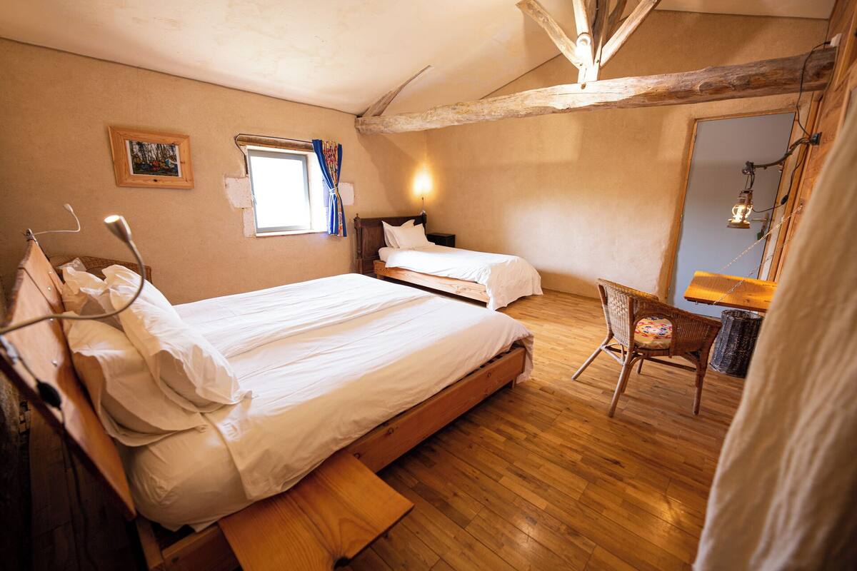 Ein Schlafzimmer mit zwei Betten, Holzboden, einem Fenster mit Vorhängen, einer Nachttischlampe und einem Korbstuhl.