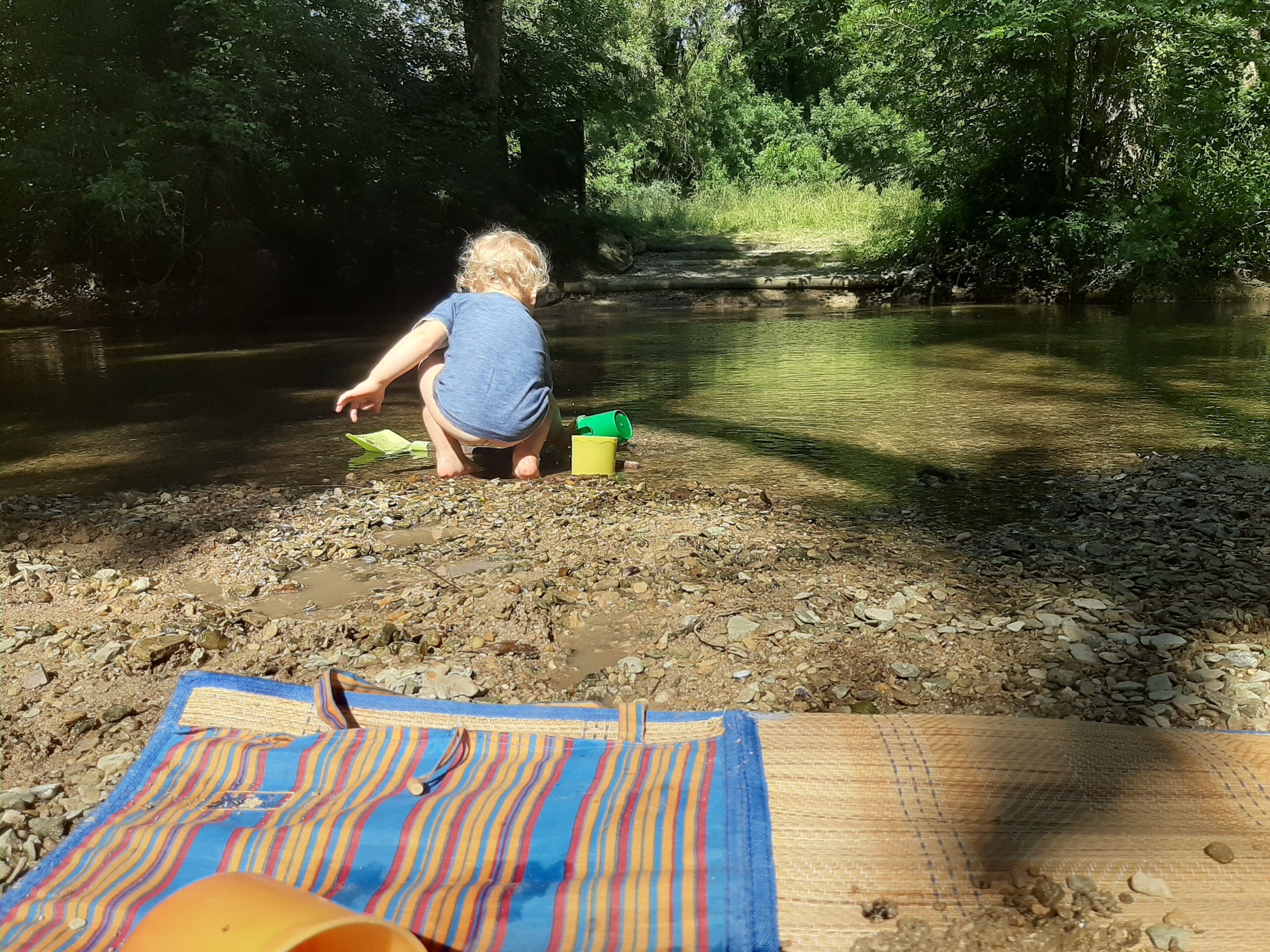A child playing by a river with a blanket and a bucket nearby.