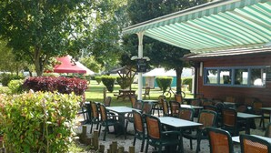 An outdoor cafe with tables and chairs, surrounded by greenery and a covered seating area. - Ridorev (Auxonne)