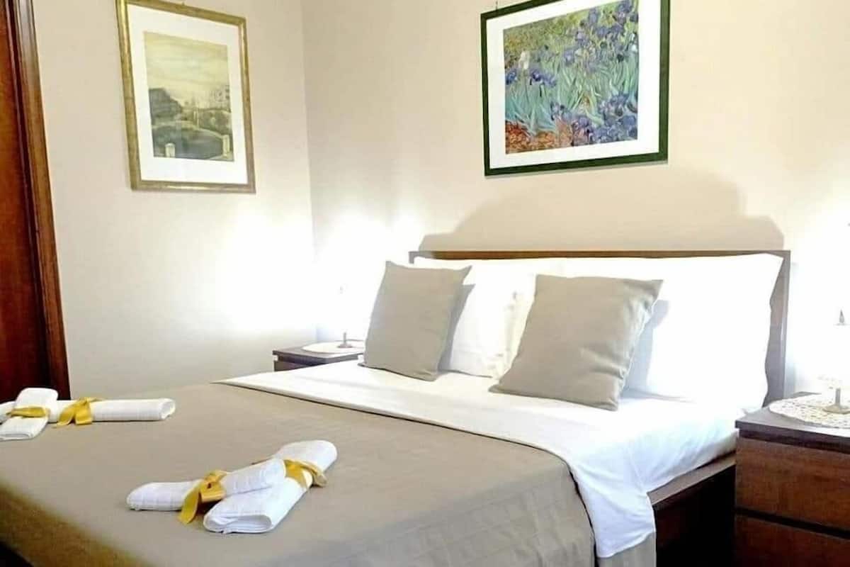 A neatly made bed with a white bedspread and pillows, a wooden nightstand with a lamp, and two framed artworks on the walls.