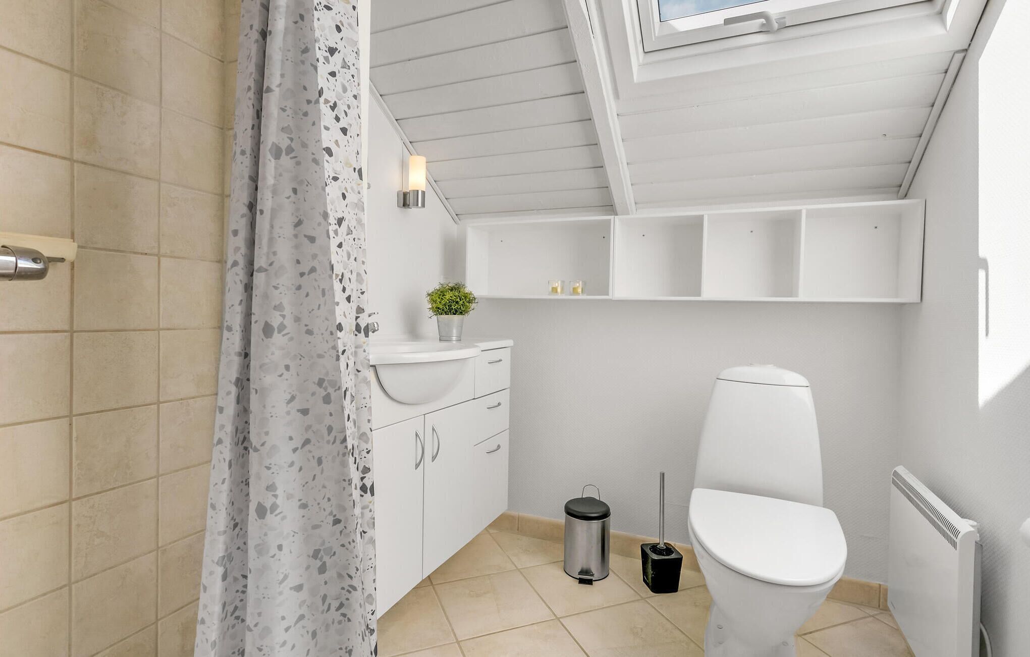 Ein kleines, modernes Badezimmer mit Toilette, Handwaschbecken und Mülleimer. Eine Deckenleuchte und ein Fenster befinden sich über der Toilette.