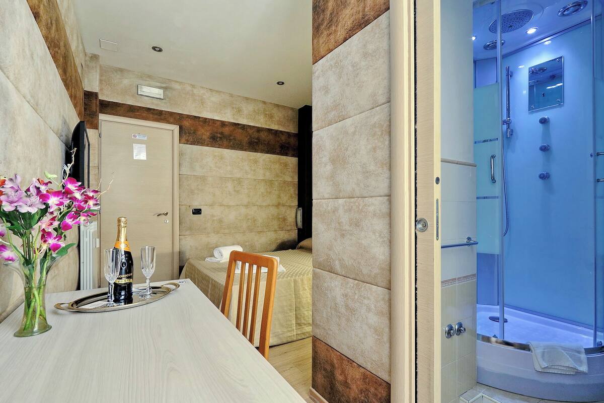 A hotel room with a bed, a shower, a dining table with a bottle of champagne and glasses, and a vase of flowers.
