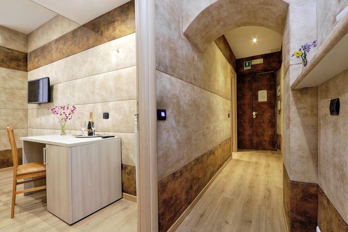 A modern hotel room with a wooden floor, a small wooden desk with a vase of flowers, a flat-screen TV mounted on the wall, and a door leading to another room.