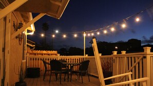An outdoor deck with a wooden railing, a table with chairs, string lights, and a potted plant. - Well-Furnished Oceanside Home! (Pismo Beach)