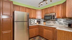 A kitchen with wooden cabinets, stainless steel appliances, a tiled backsplash, and a colorful rug. - Oceanfront Remodeled Studio Condo (Kailua-Kona)