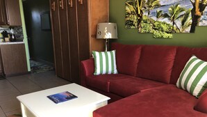 A living room with a red sofa, a white coffee table, and a green and white striped pillow. There is a painting of a tropical scene on the wall. - Oceanfront Remodeled Studio Condo (Kailua-Kona)