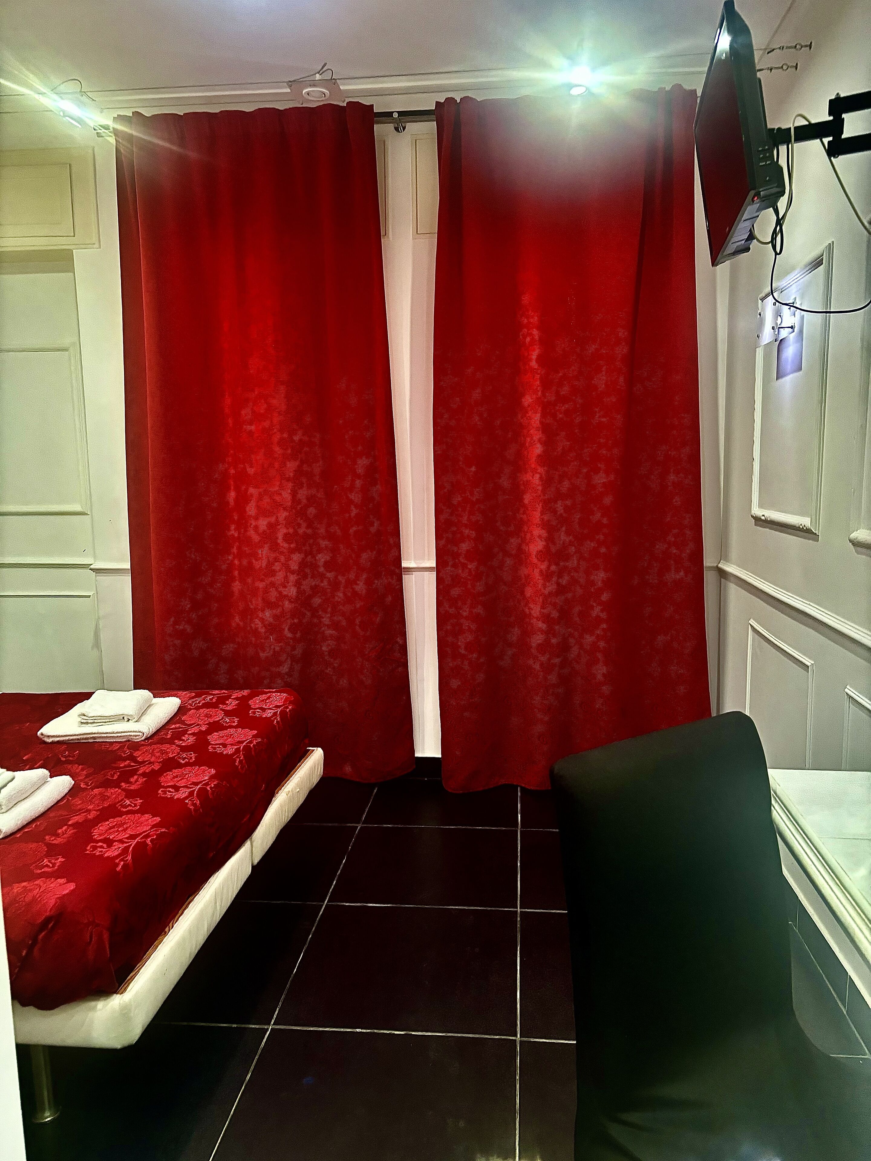 Una habitación con dos camas, cortinas rojas, una silla negra y un televisor montado en la pared.